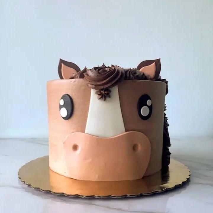 A cake decorated to look like a horse's head. The cake is a light brown color with a dark brown mane and nose, along with two pointy ears.