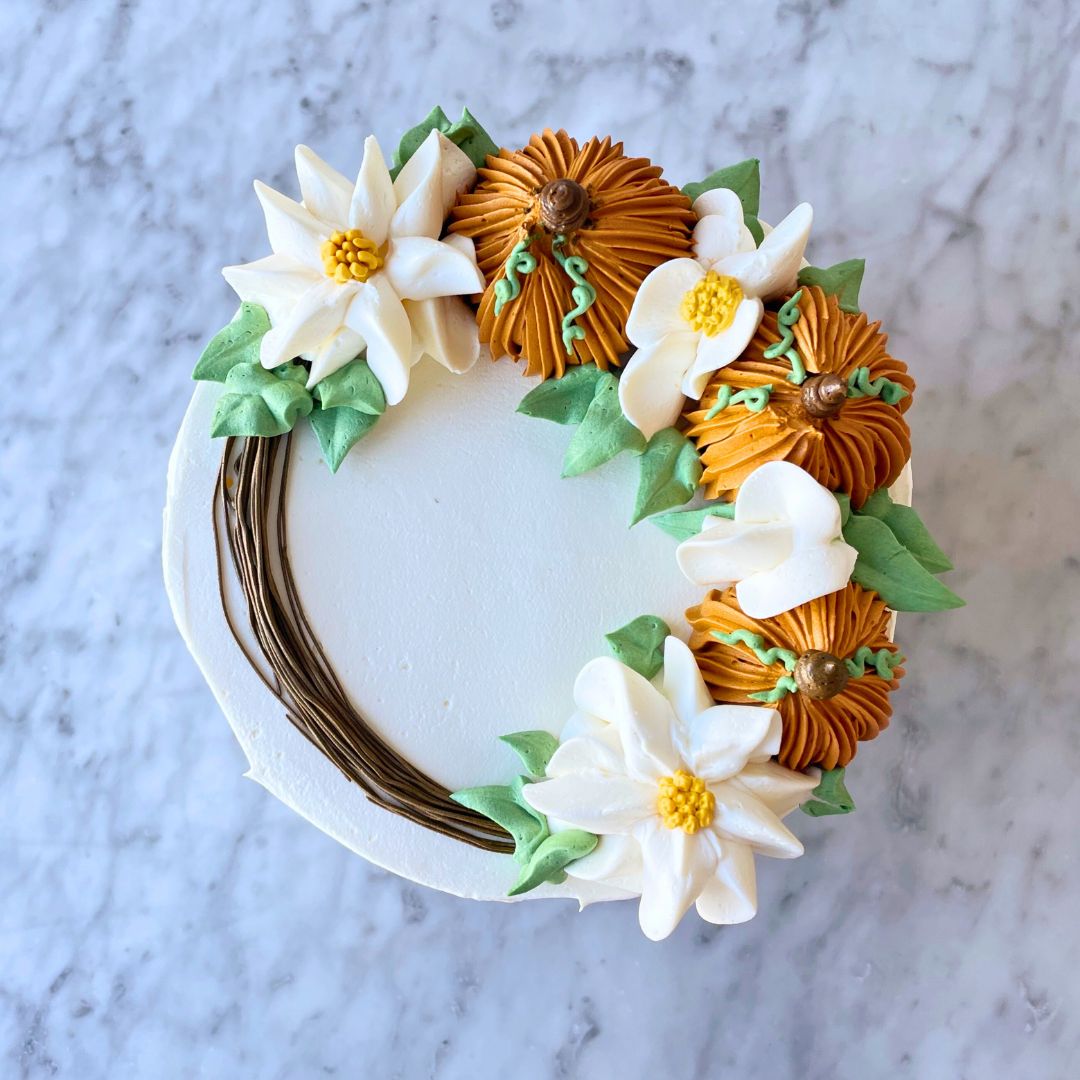 An overhead view of a cake decorated with a wreath of buttercream flowers, pumpkins, and leaves.