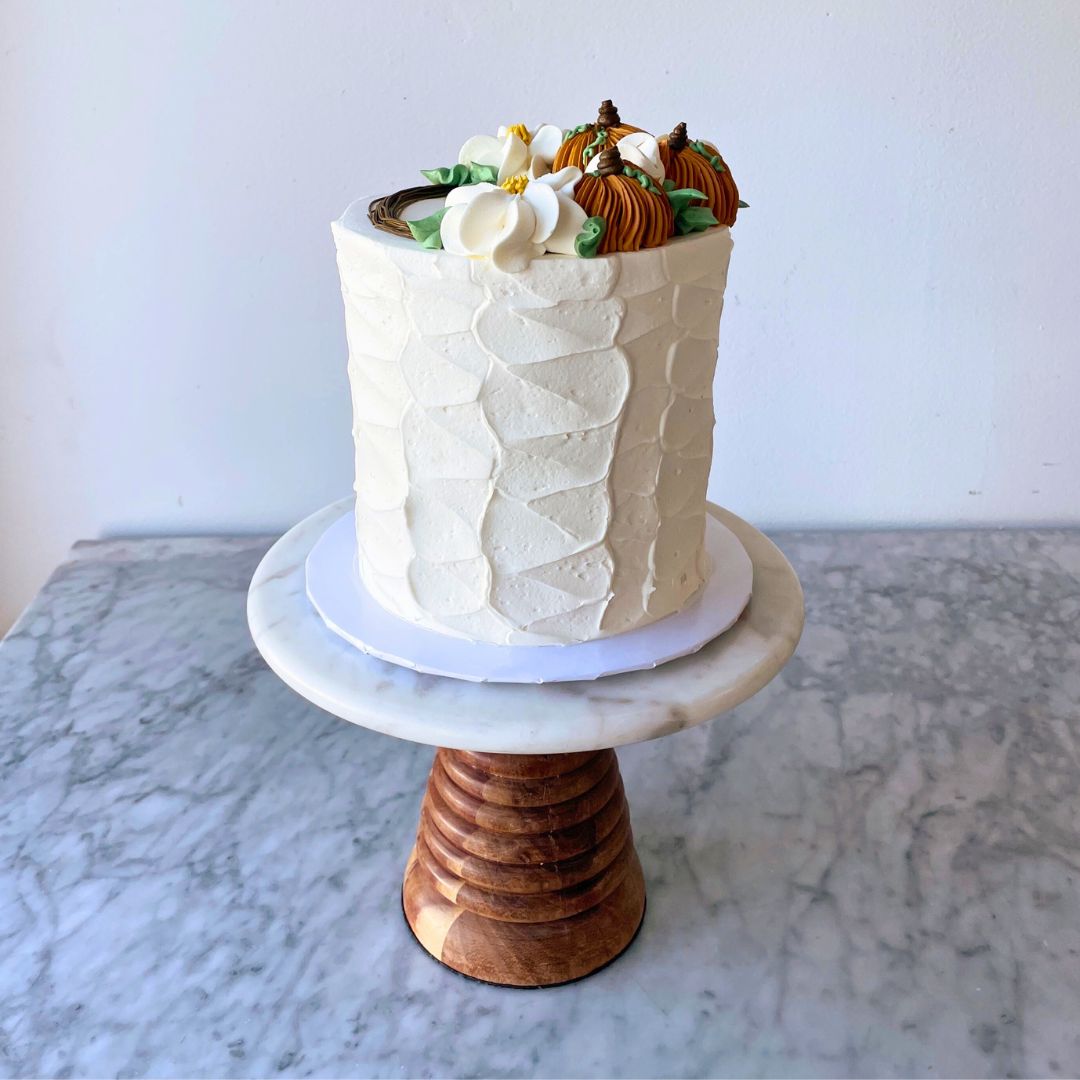 Two-tier tall small round white cake with buttercream flowers and orange pumpkins designs on top.