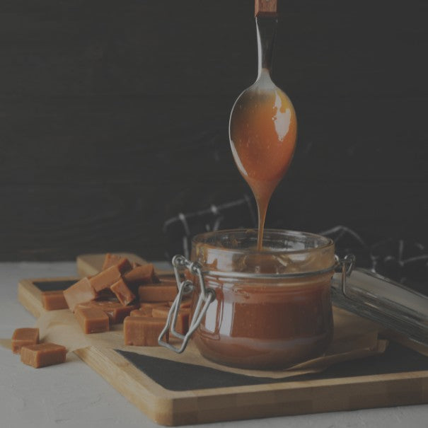 Jar of caramel sauce with a spoon dripping onto a wooden board with caramels.