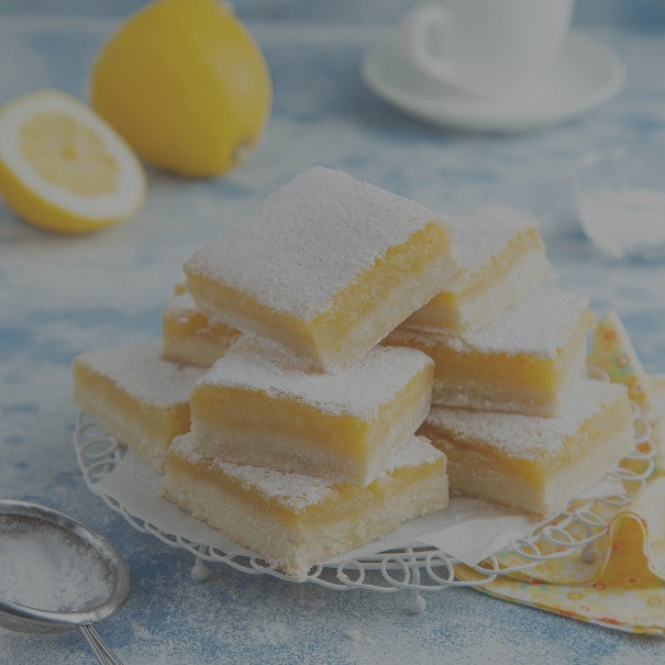 Stack of lemon bars on a decorative plate with lemons and a teacup in the background.