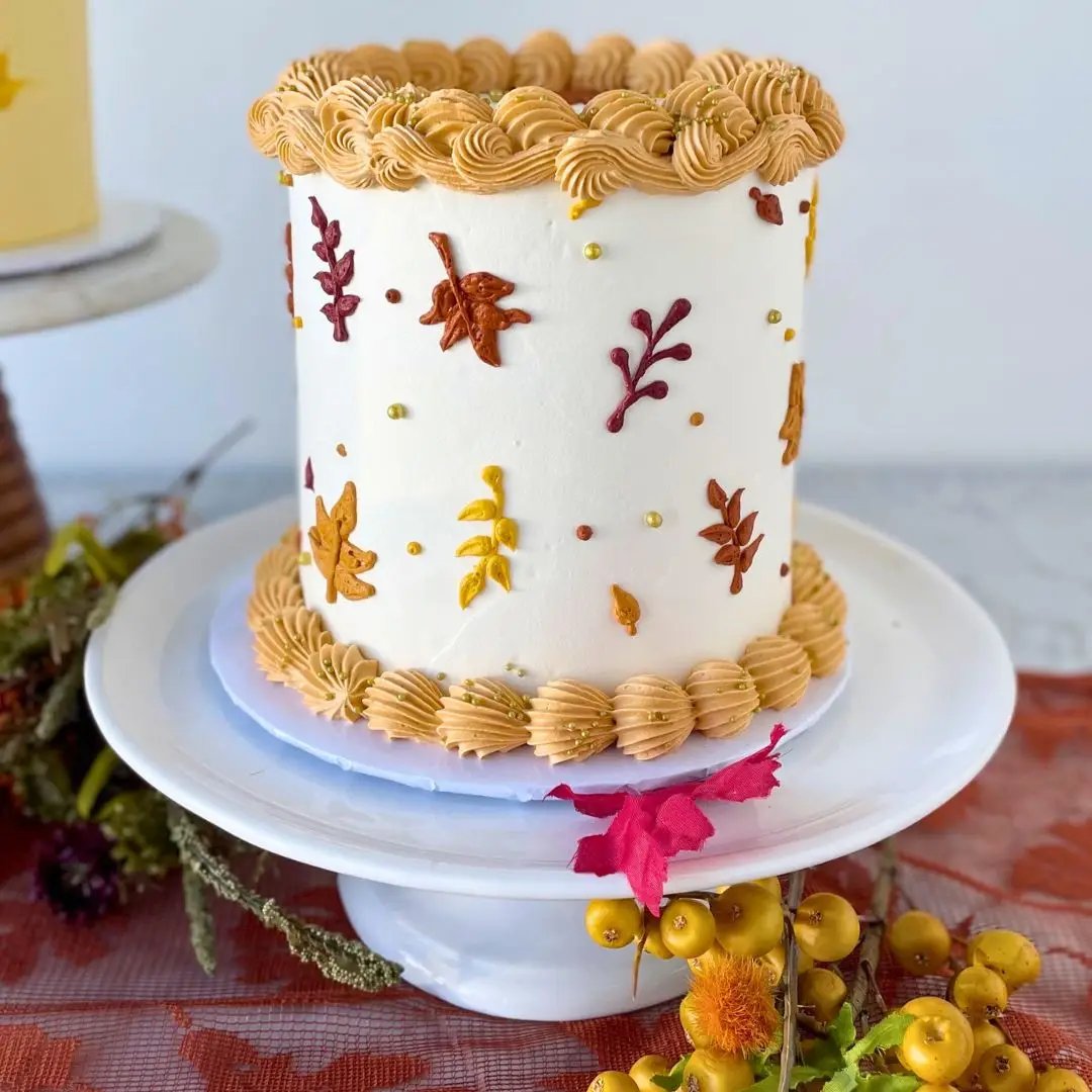 A two-tier small round cake with ridged piping on the top and bottom, and a design of colorful autumn leaves, on a white cake stand.