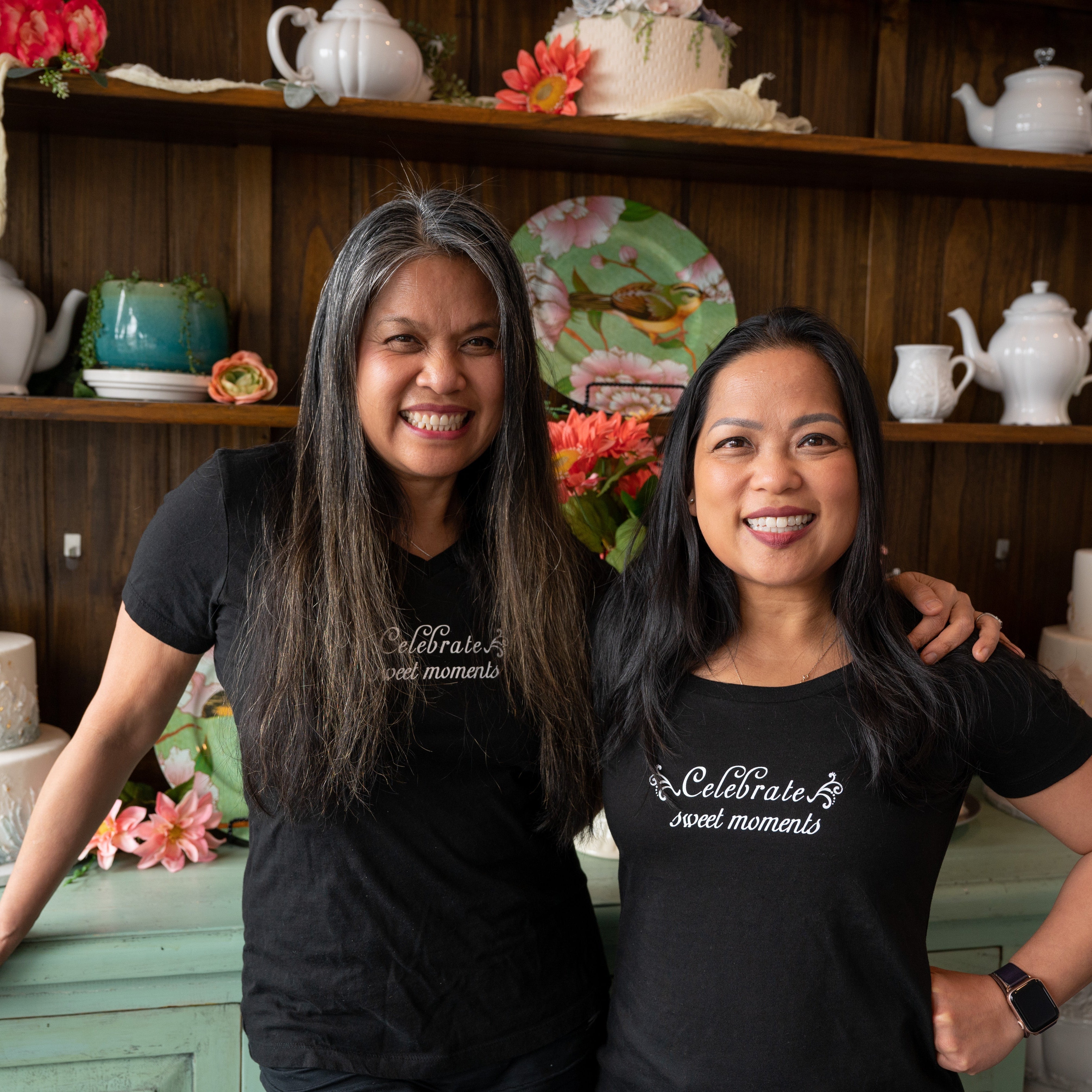 Two women wearing black branded t-shirts smiling and posing side by side in a bakery or café setting, both appearing cheerful and friendly