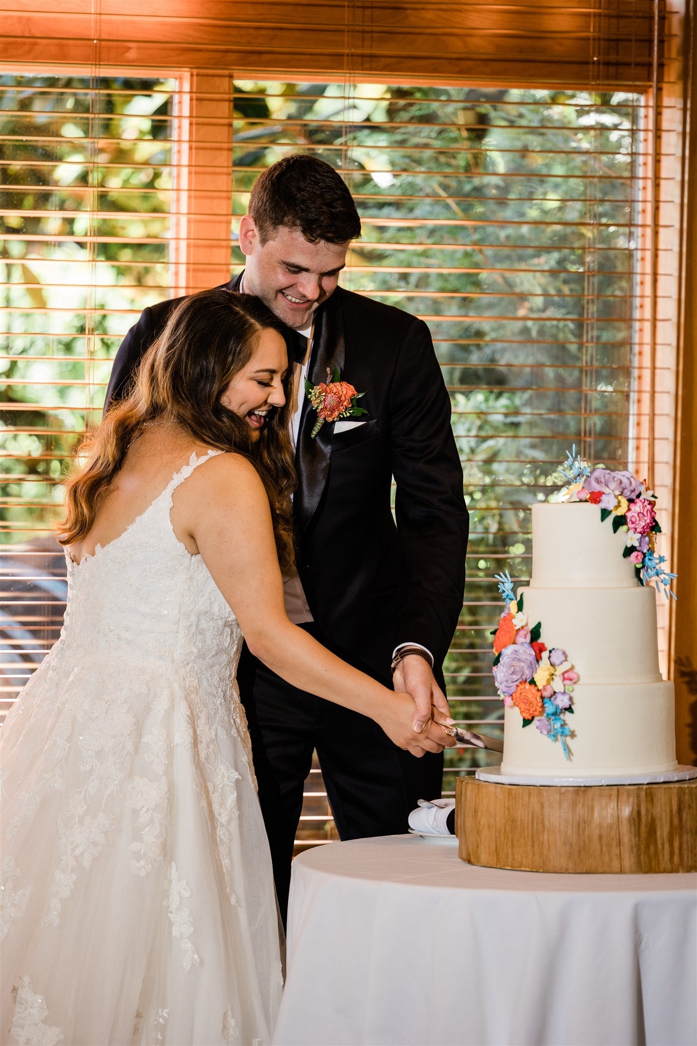Bride and groom sharing a kiss while cutting their wedding cake together in an indoor venue decorated with greenery and floral arrangements