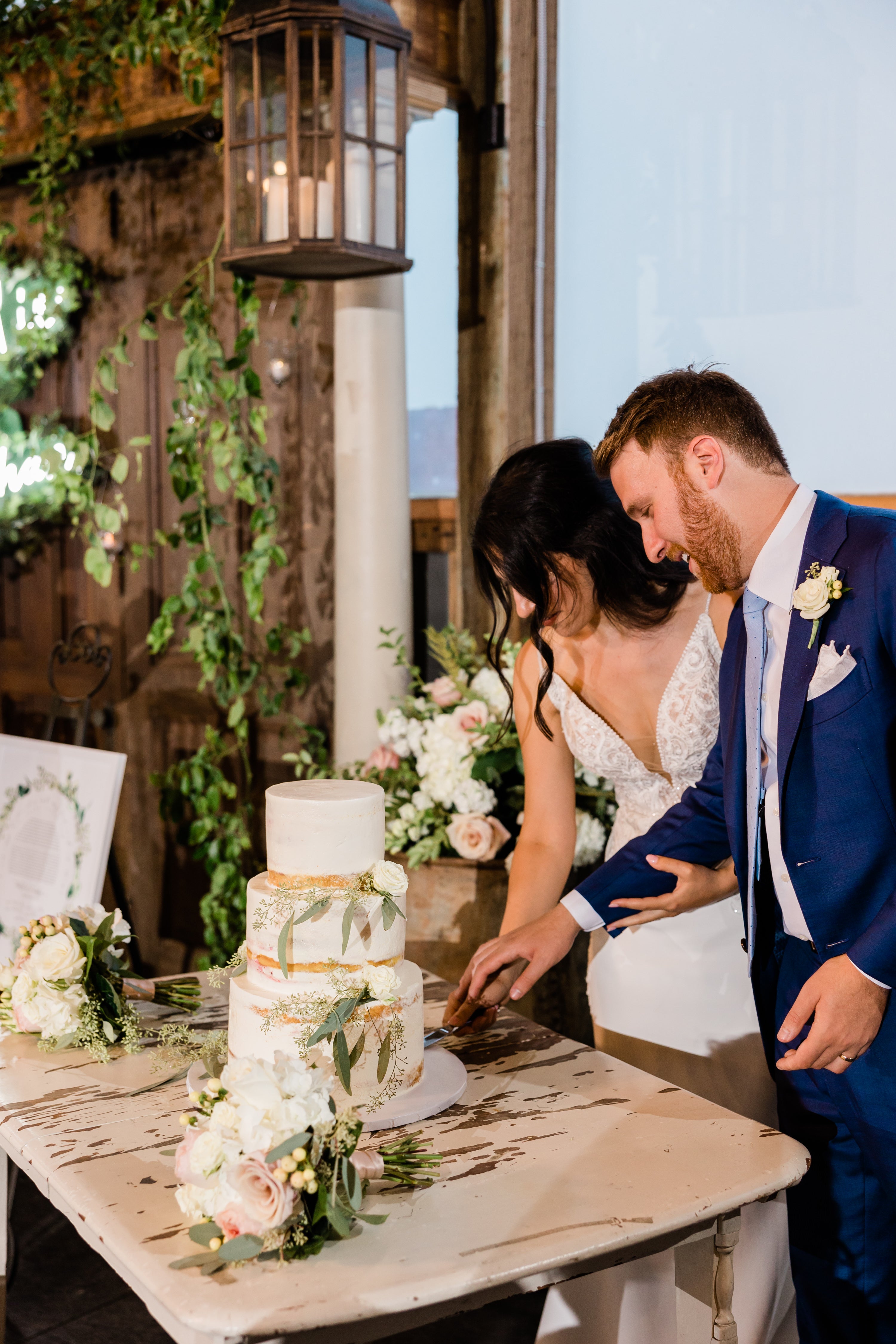 Bride and groom cutting a three-tier white wedding cake decorated with greenery and soft blush roses