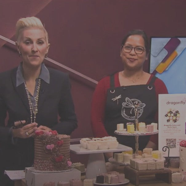 Two women standing behind a display of cakes and pastries in a bakery setting.
