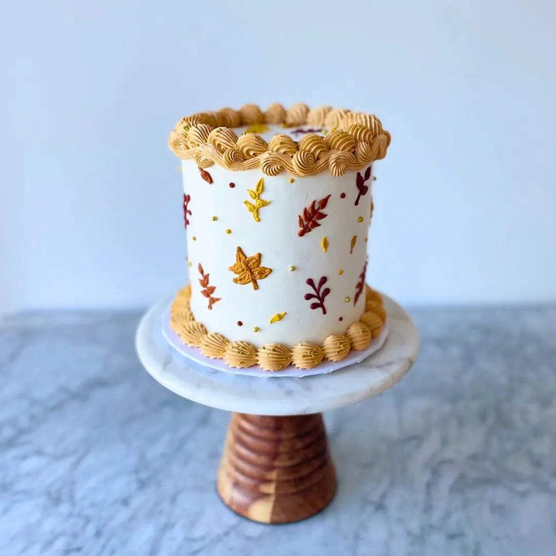 Two-tier small round cake with ridged piping on the top and bottom, and a design of colorful autumn leaves, on a wooden cake stand.