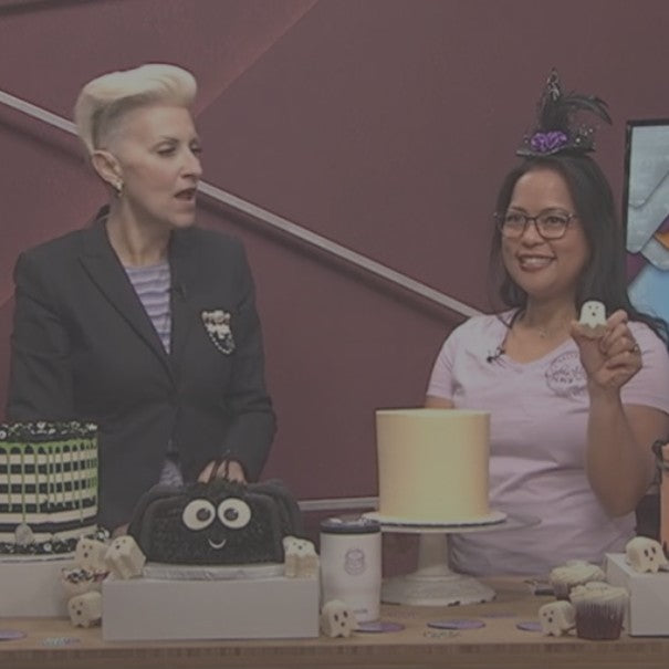 Two women standing behind a table with cakes and small figures, one holding a piece of cake.