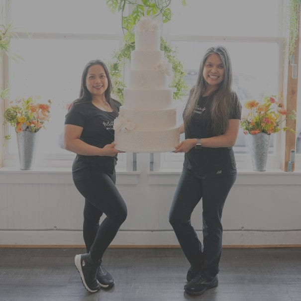 Two women holding a large wedding cake in a room with floral decorations.