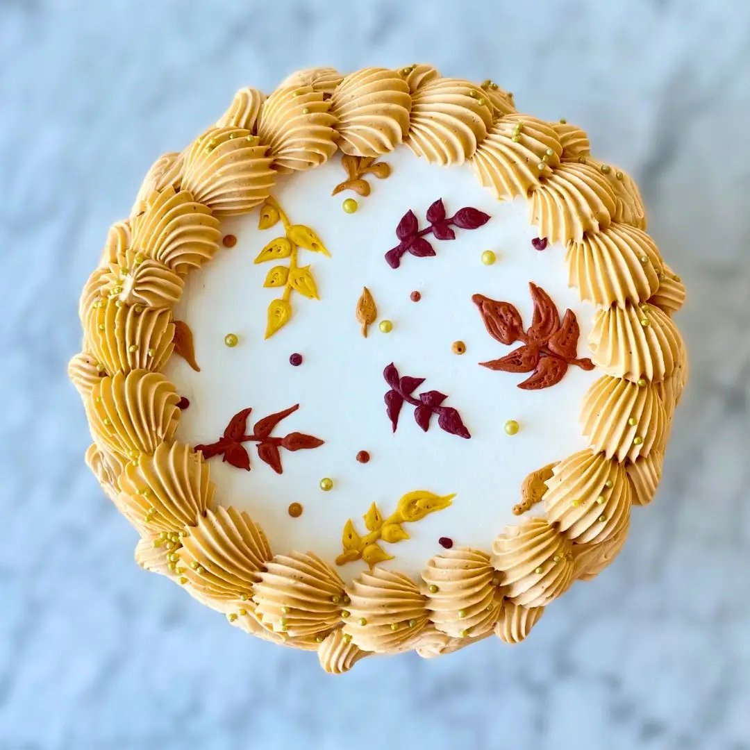 An overhead view of a small round cake with a piped ridged border and a design of colorful autumn leaves on top.