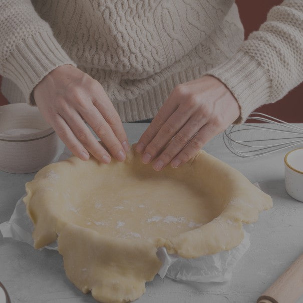Person making a pie crust on a kitchen counter with various baking tools.