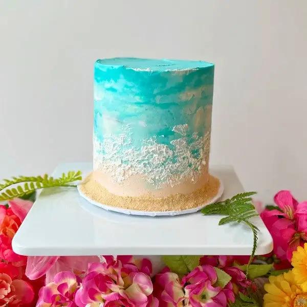 A beach-inspired chocolate raspberry cake with blue-tinted buttercream "waves", and white "sand", placed on a white surface with colorful flowers around it.