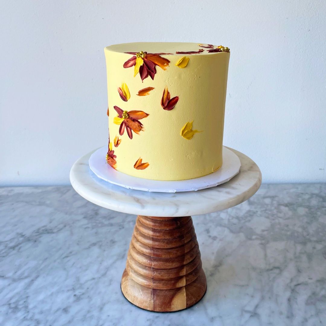 A tall round cake with pale yellow frosting, decorated with small painted autumn leaves in shades of red, orange, and yellow.
