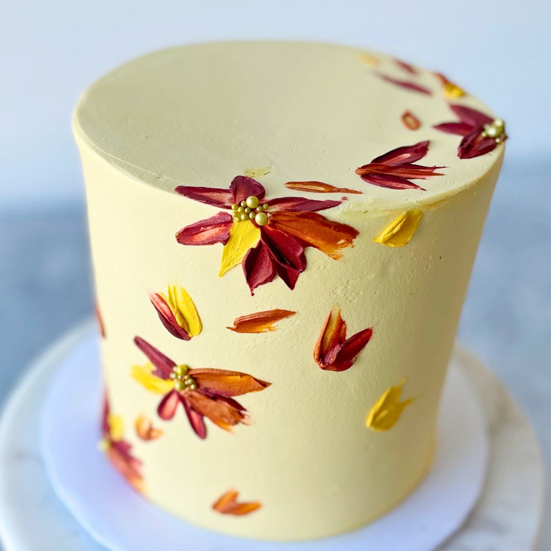 Two-tier tall round cake with pale yellow frosting, decorated with small painted autumn leaves in shades of red, orange, and yellow.