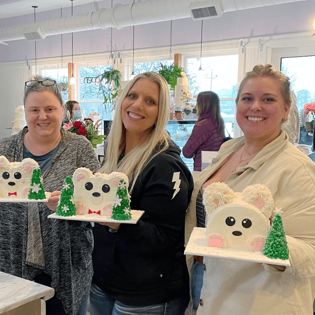 Three women stand side by side, smiling at the camera and holding up their completed polar bear-shaped cakes decorated with green, tree-shaped icing.