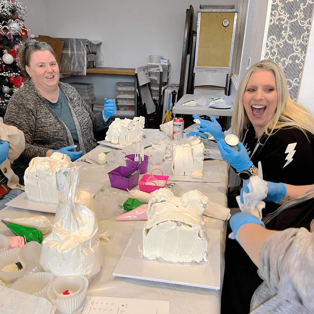 Two women laughing while decorating white cakes in the shape of animals at a table.