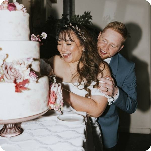 A bride and groom smiling while posing behind their tiered wedding cake