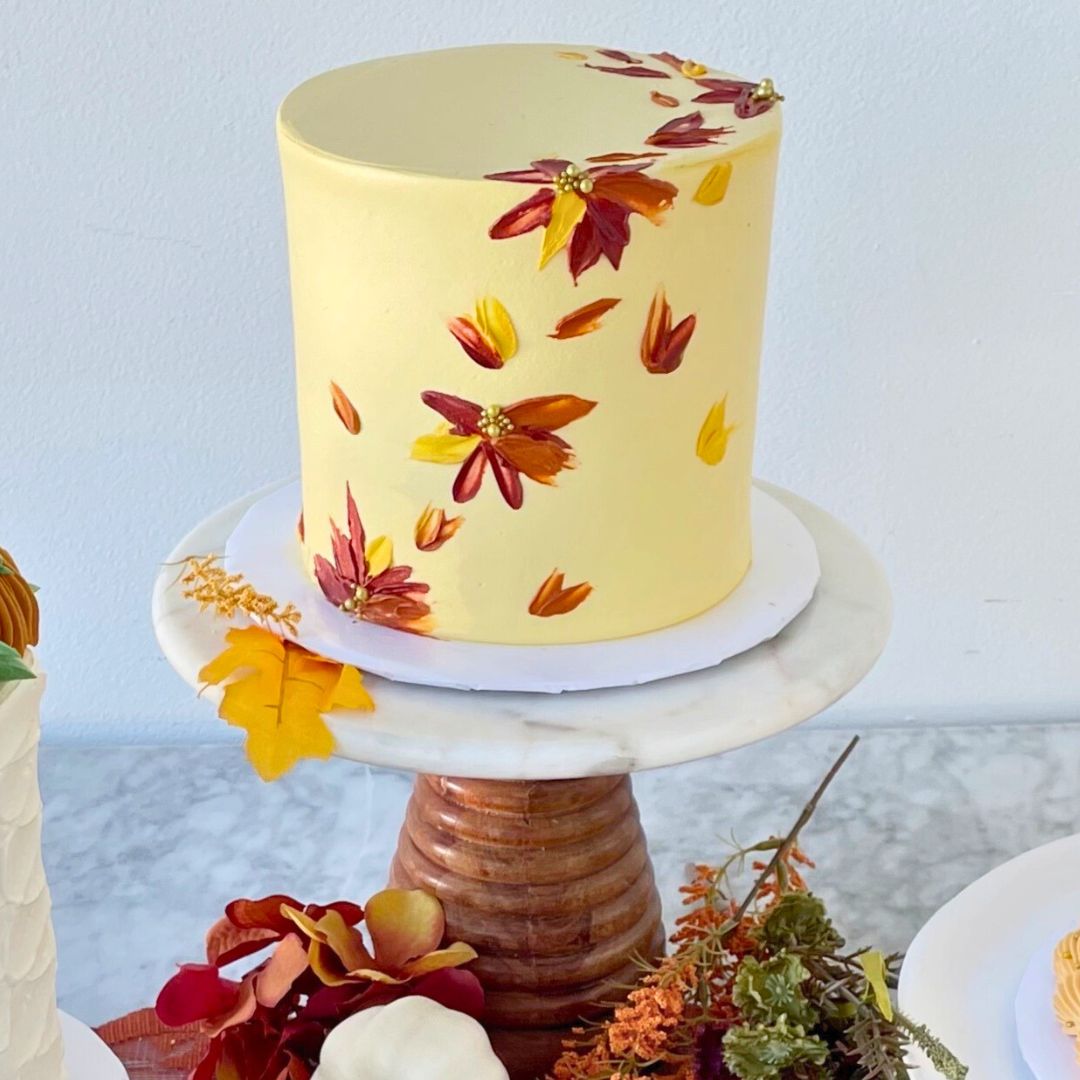 Two-tier tall round cake with pale yellow frosting, decorated with small painted autumn leaves in shades of red, orange, and yellow.