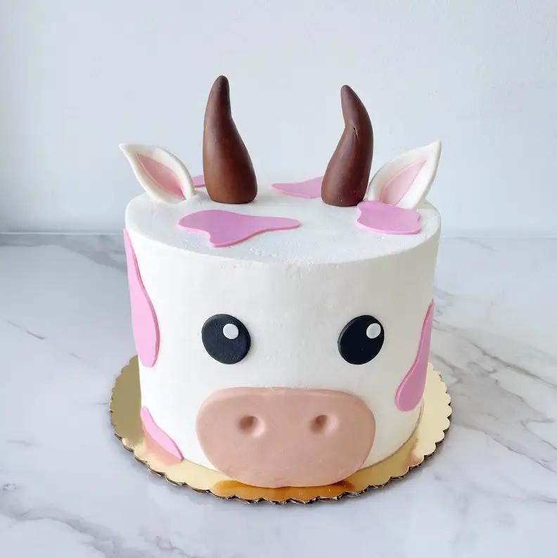 A round, white cake decorated to look like a cow's head. The cake has large pink splotches, a pink nose, two dark brown horns, and pointy ears.
