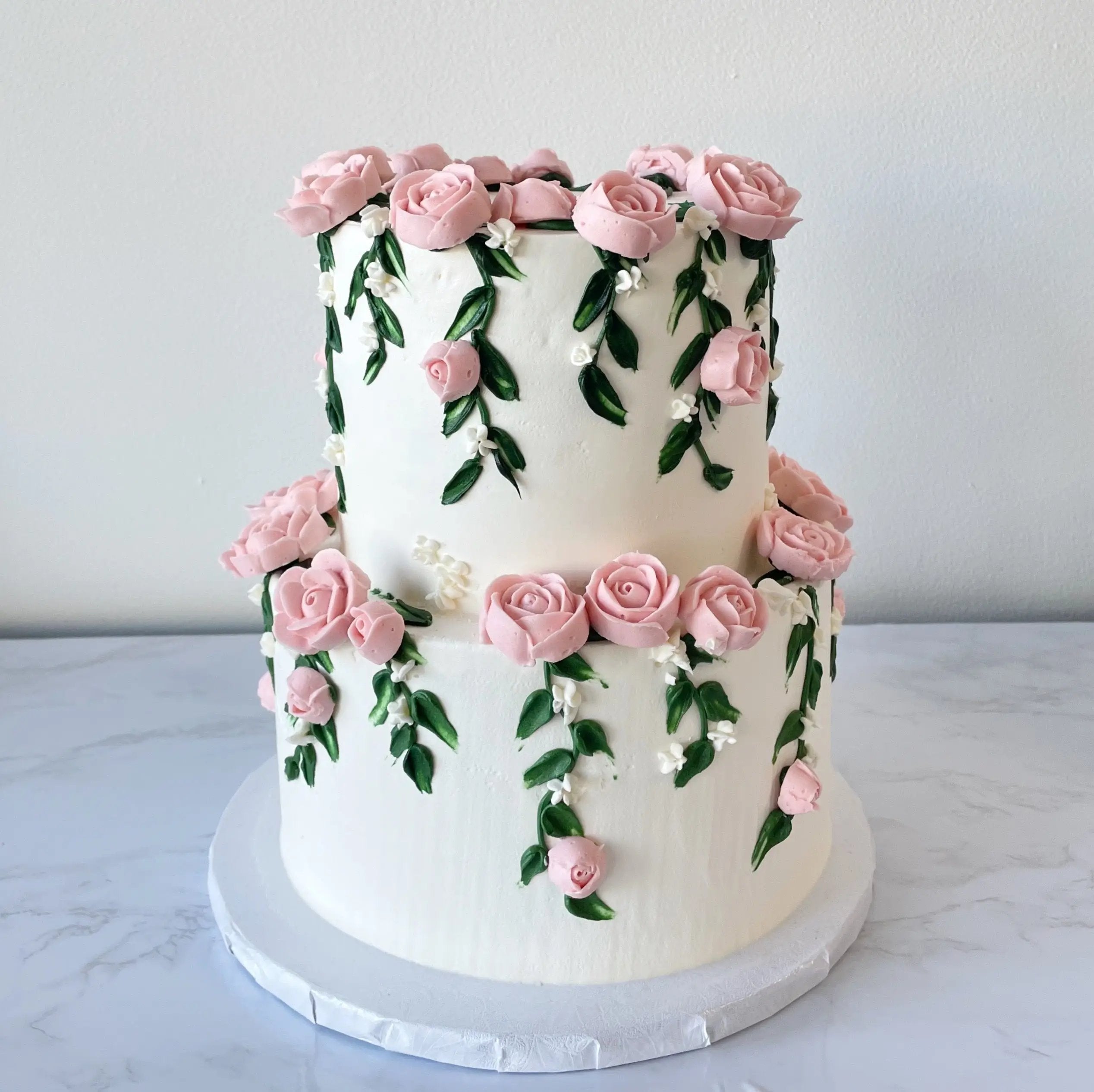 A two-tiered white cake with piped pink flowers and green vines on the side.