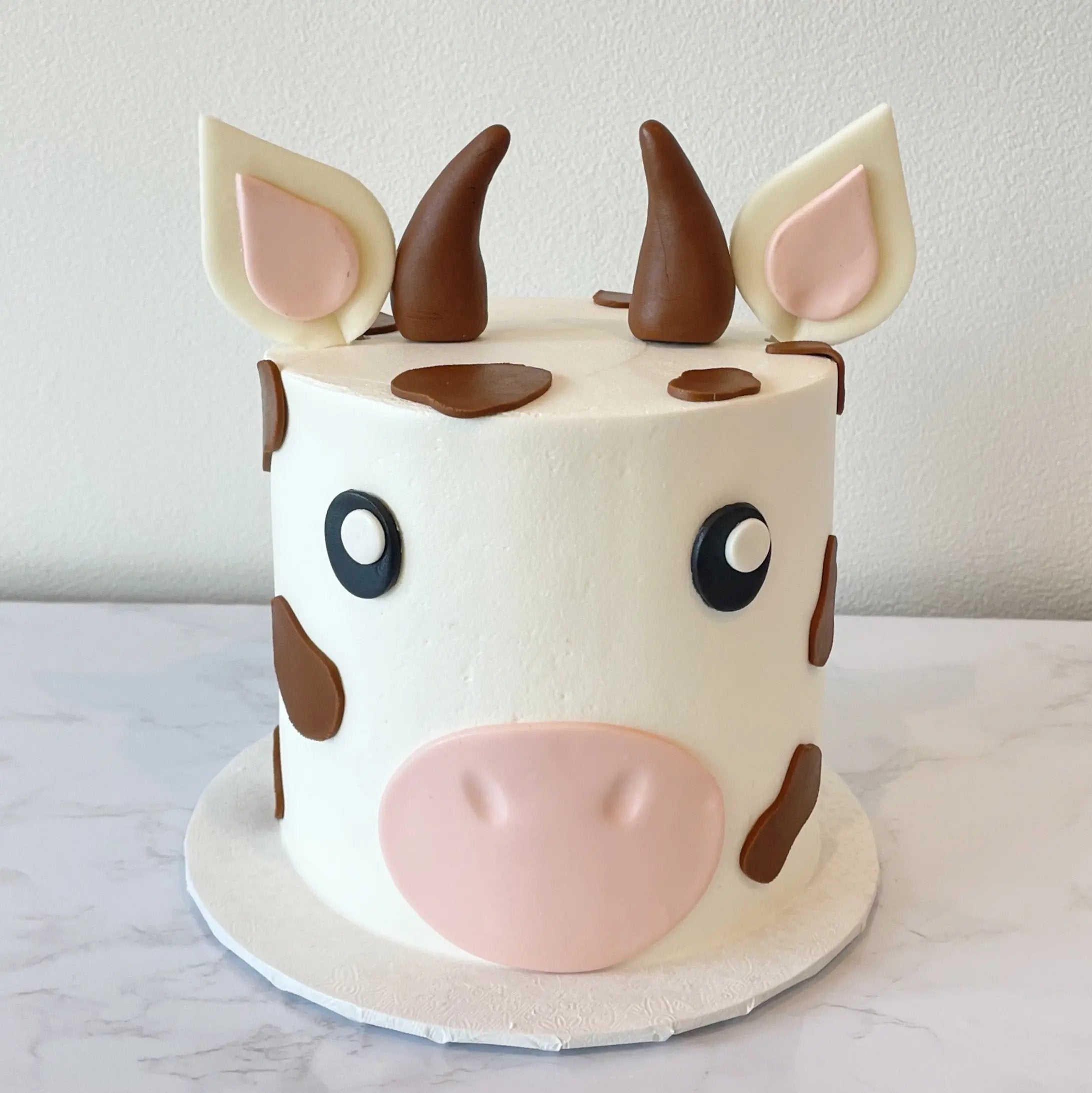 A single-tiered cake decorated to look like a brown-and-white cow's face, with pink ears, chocolate horns, and black eyes.