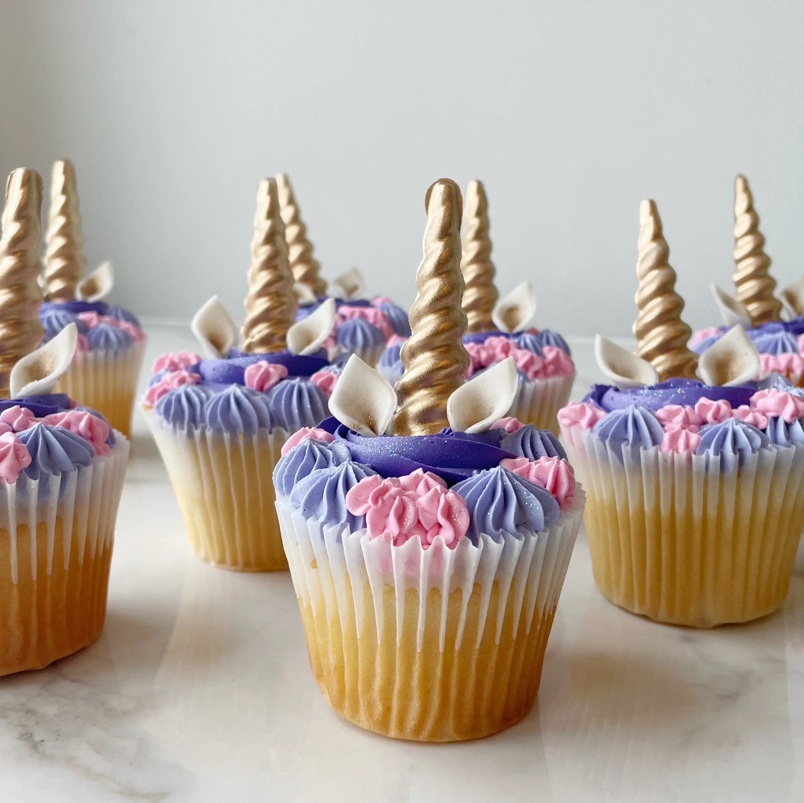 A group of cupcakes with purple, blue, and yellow frosting, each topped with a gold unicorn horn.