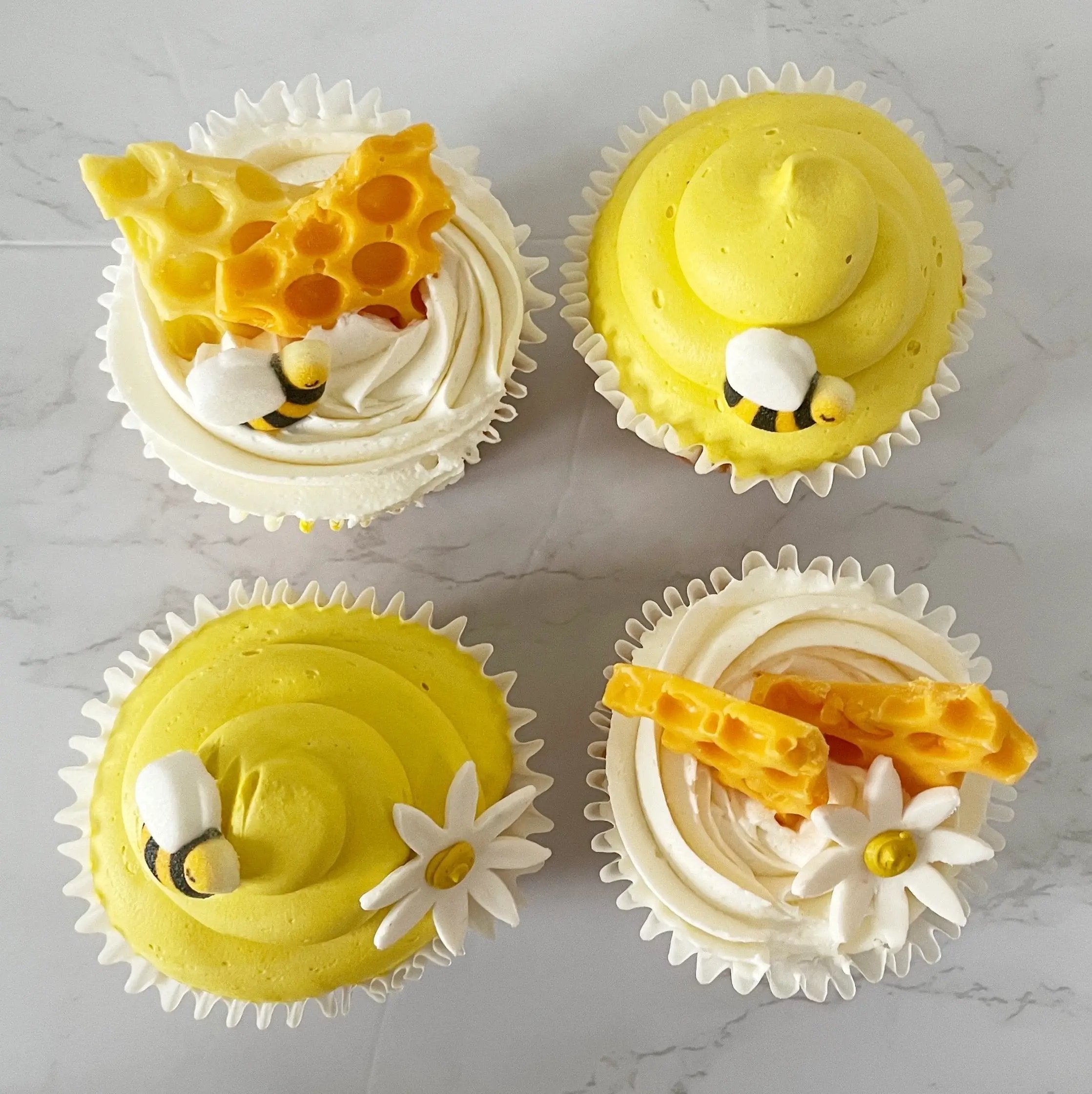 A top-down view of four cupcakes with yellow and white frosting, decorated with small flowers and bees.
