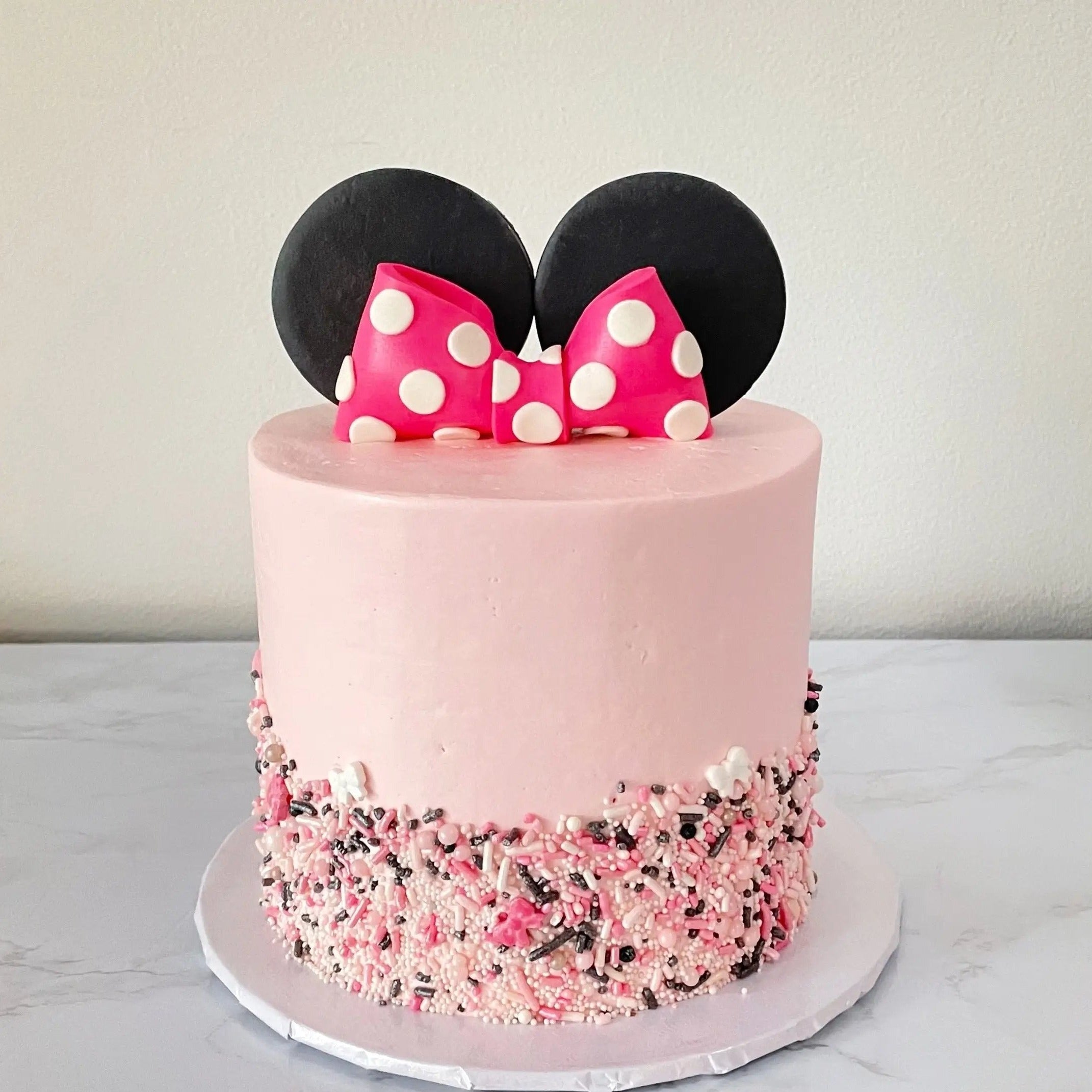 A small, pink Minnie Mouse-themed cake with black ears and a pink bow with white polka dots on top. The bottom of the cake is covered in pink, white, and black sprinkles.