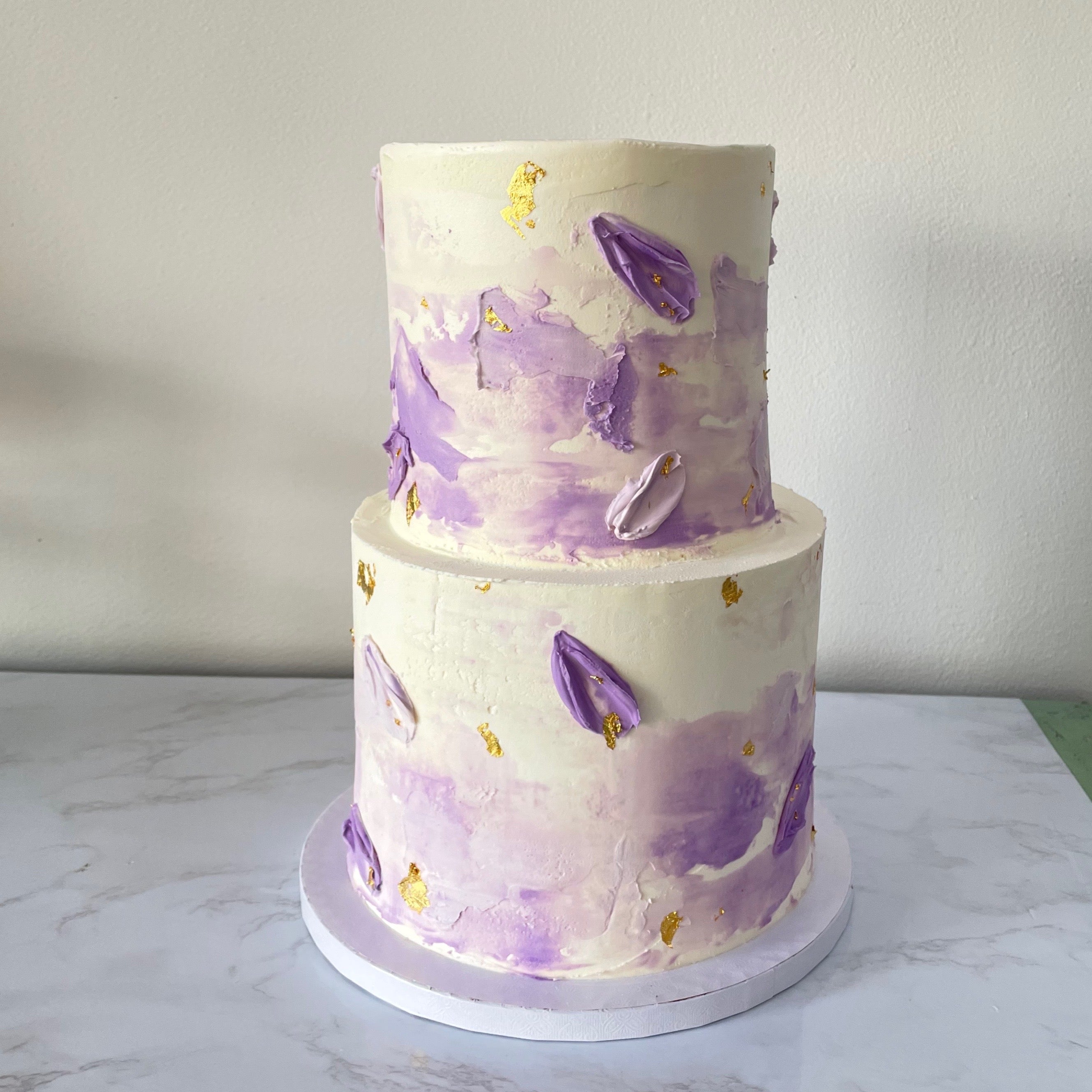 A two-tiered cake with white frosting and abstract purple and gold leaf decorations