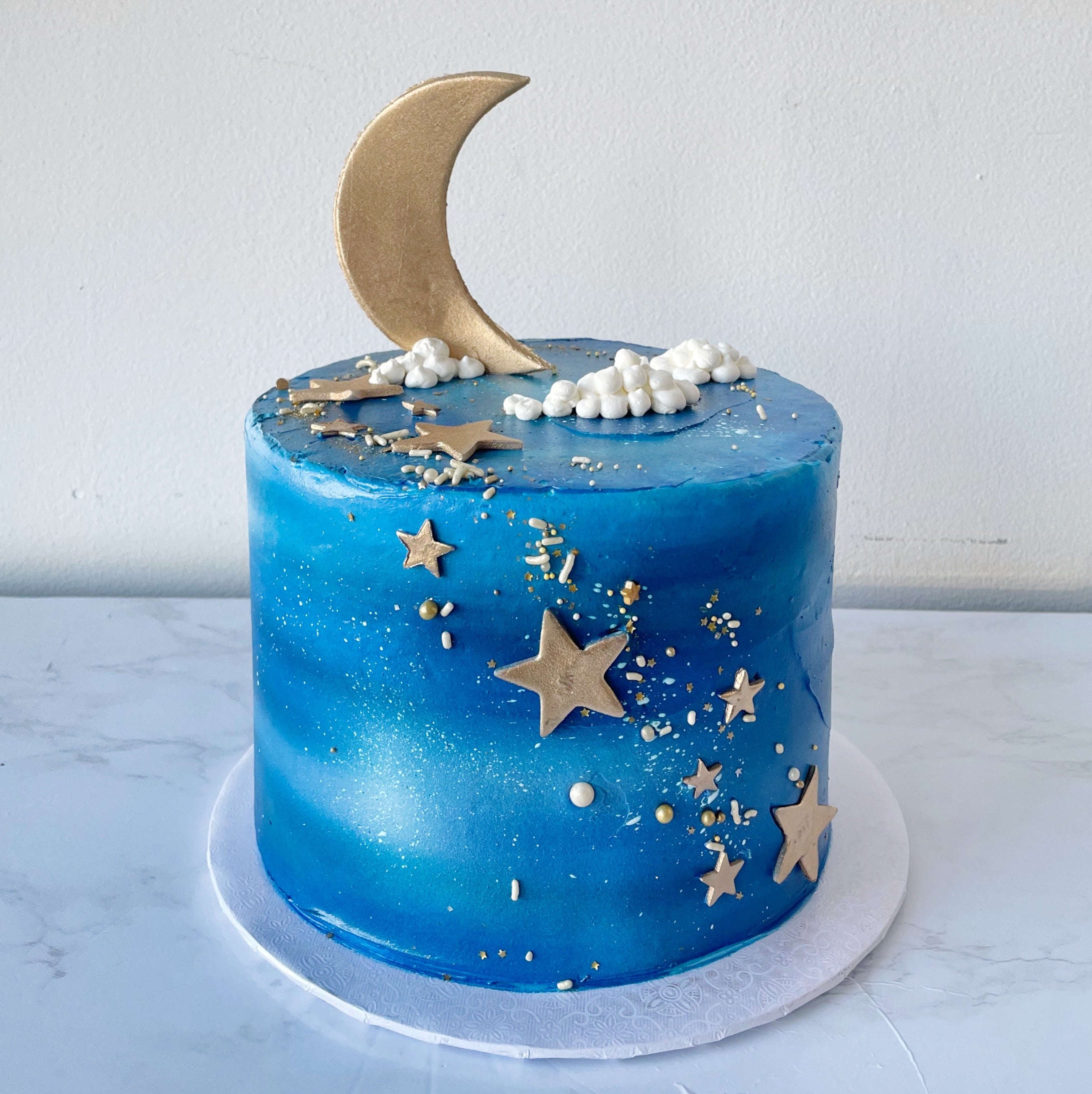 A round, blue cake decorated with a gold crescent moon, stars, and clouds