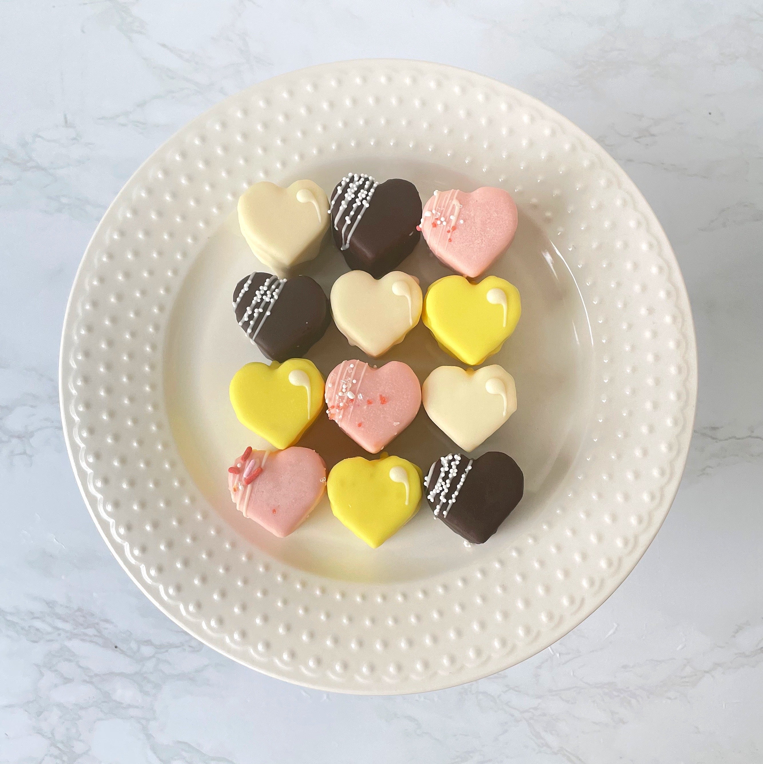A dozen heart-shaped chocolates in white, yellow, pink, and dark chocolate on a white plate