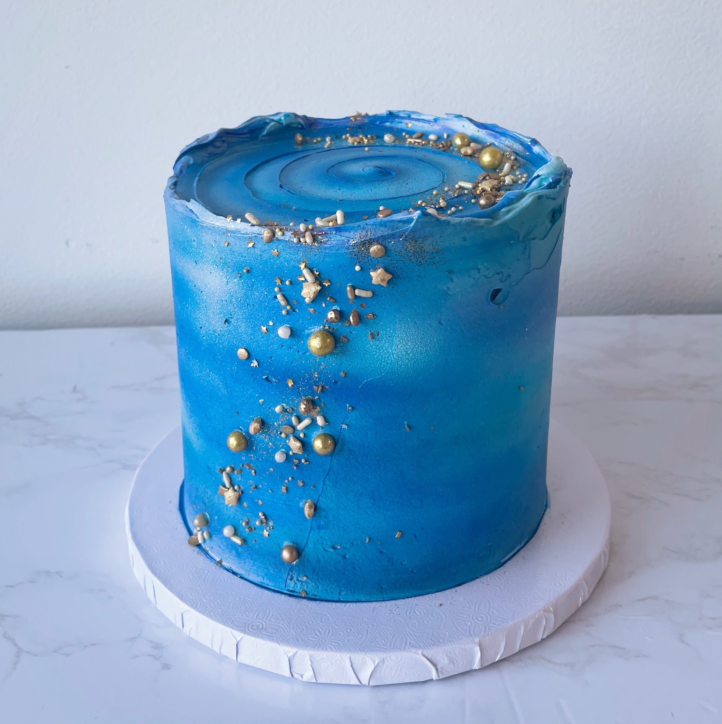 A small round cake in galaxy blue with gold star decorations, placed on a white base