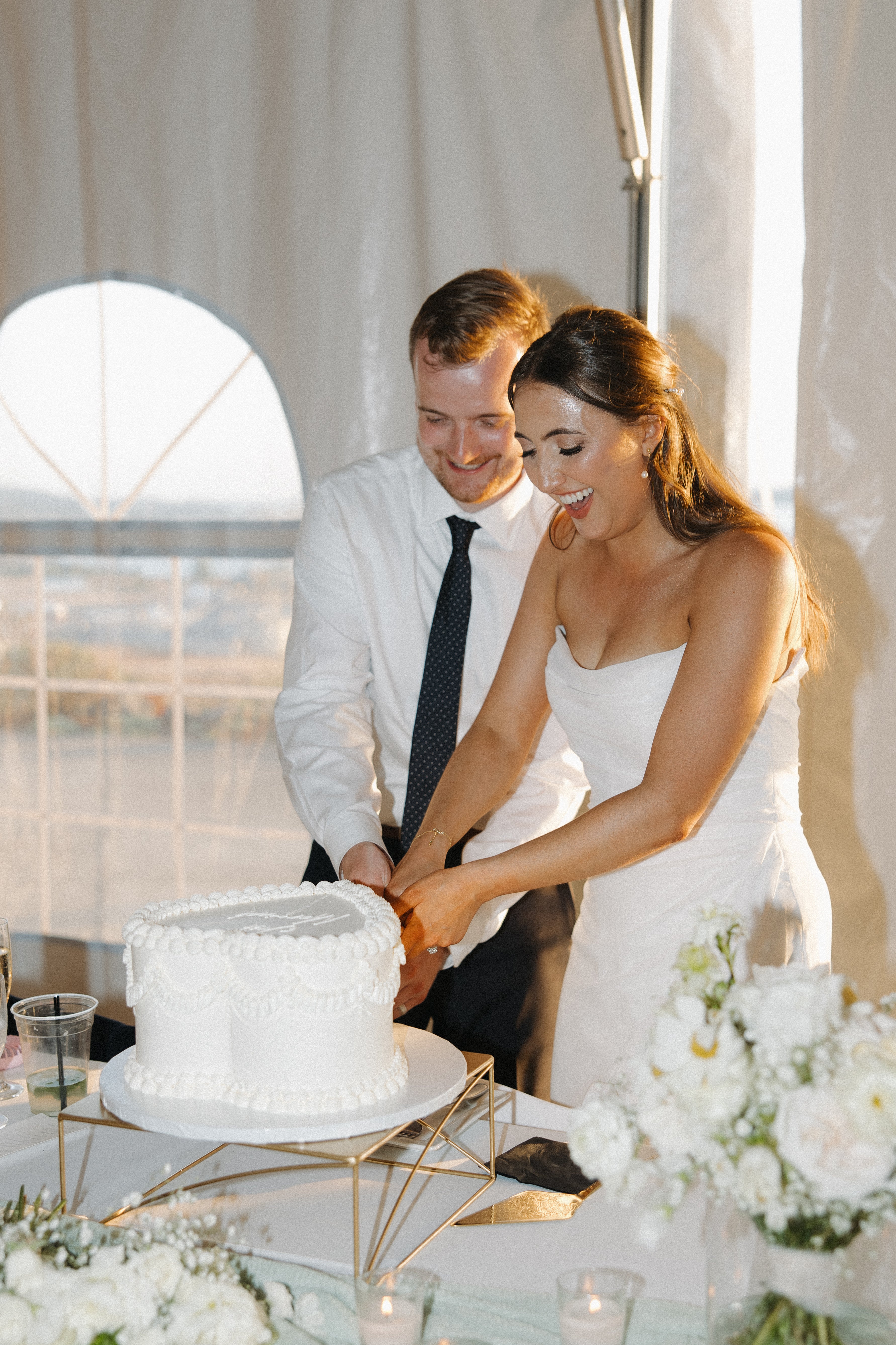 Happy couple standing close together while preparing to cut their wedding cake, with floral decor and neutral tones in the background