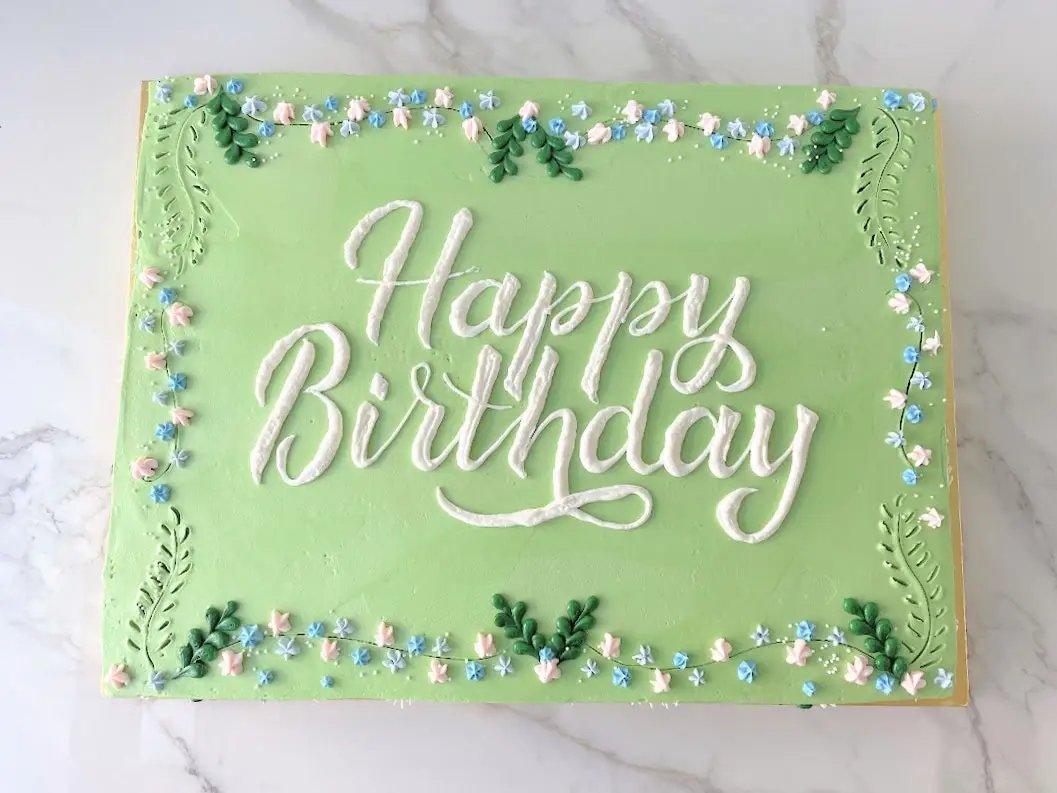 A light green rectangular cake. The words "Happy Birthday" are written in white frosting, and the edges are decorated with white frosting swirls and small blue flowers.