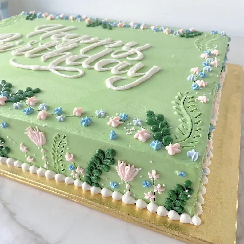 A rectangular, light green cake. The words "Happy Birthday" are written in white frosting in the center. The edges are decorated with white frosting swirls and small blue flowers.