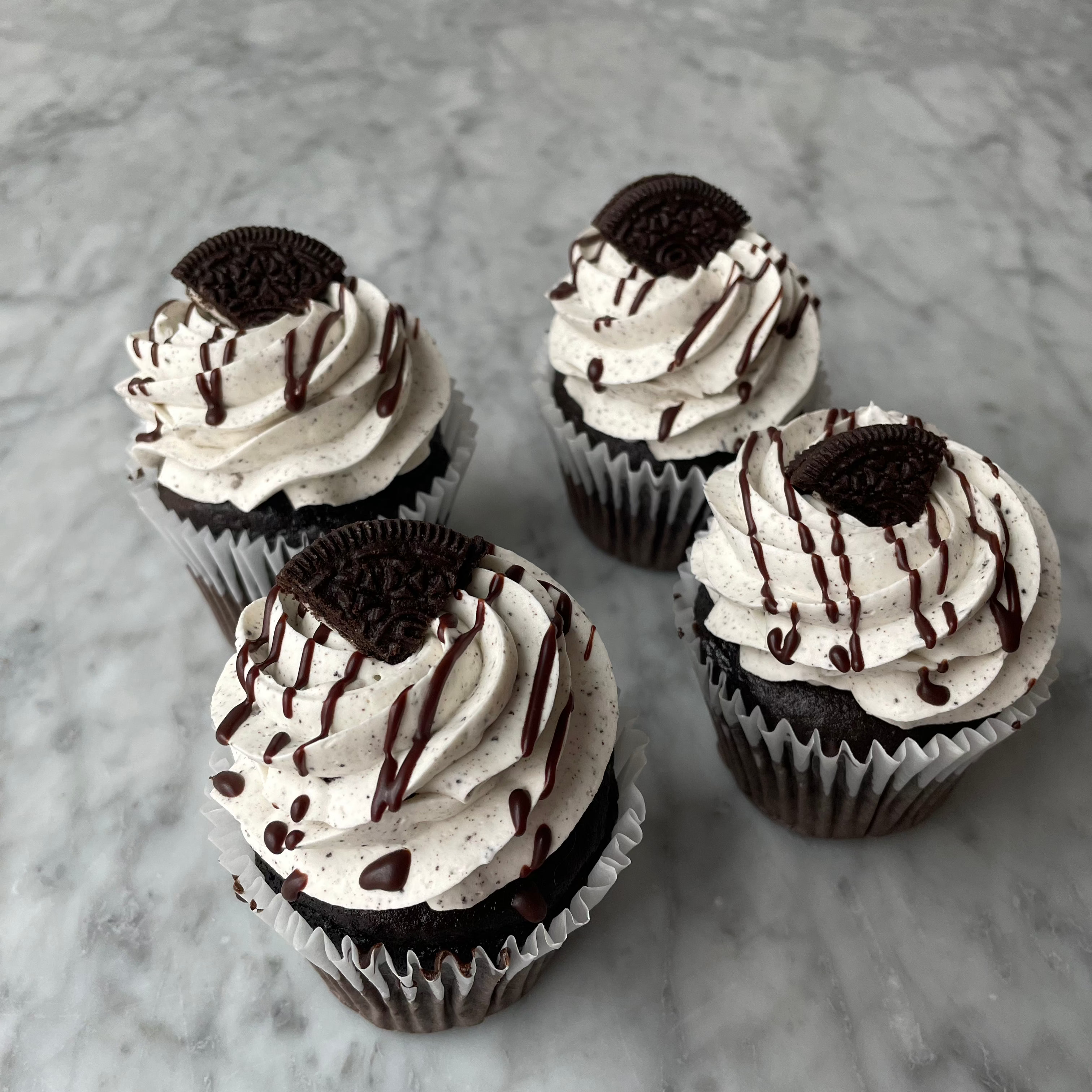 A set of four Cookies & Cream Cupcakes with chocolate cake and creamy buttercream frosting, garnished with a drizzle of ganache and a cookie piece on top, placed on a marble surface.