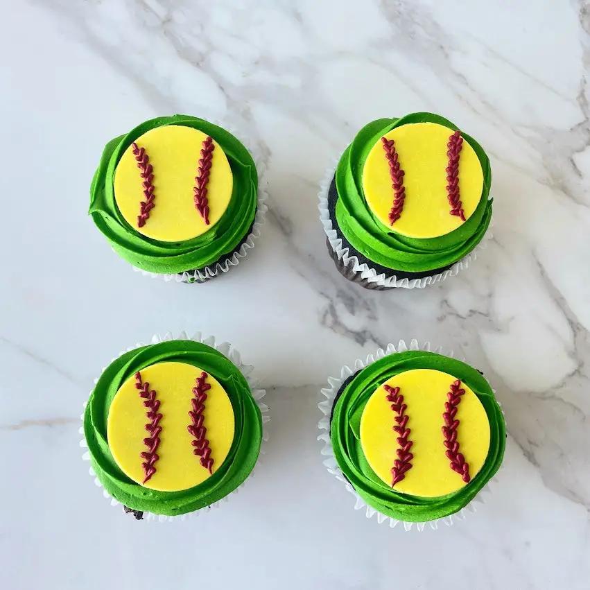 Four cupcakes with yellow frosting. Each one is decorated with two red lines to resemble a softball.