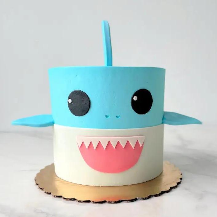 A white and light blue cake with a large shark fin on top. The cake is decorated to look like a cartoon shark's head, with a pink mouth showing a white row of jagged teeth and two large eyes.