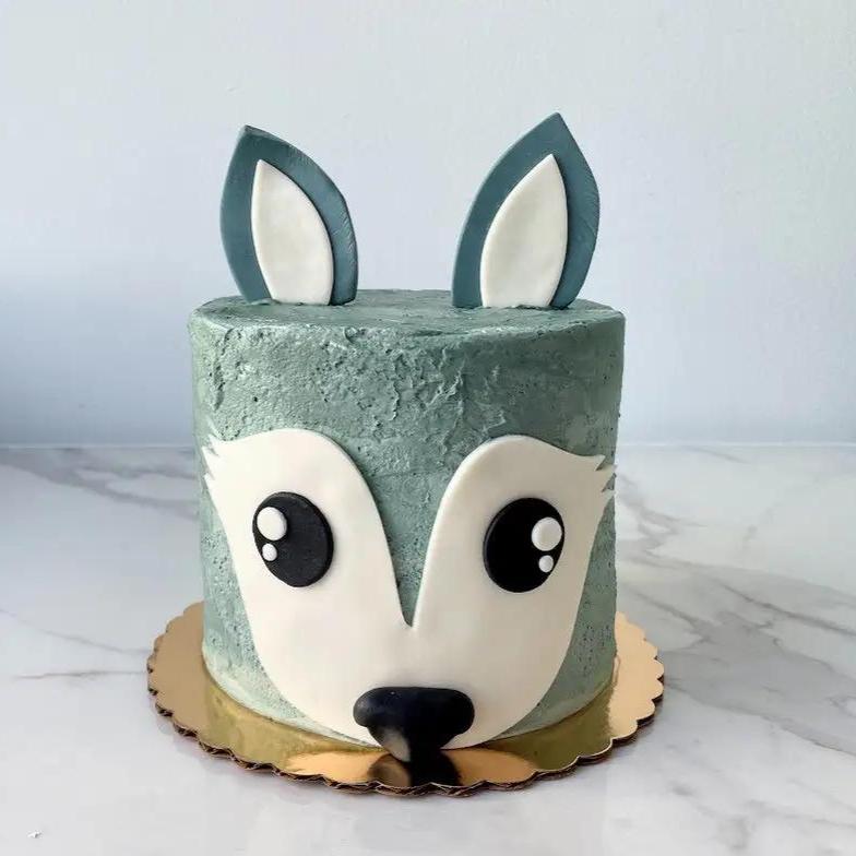 A cake decorated to look like a fox's face. The top half is gray with pointed ears, and the bottom is white with a black nose and eyes.