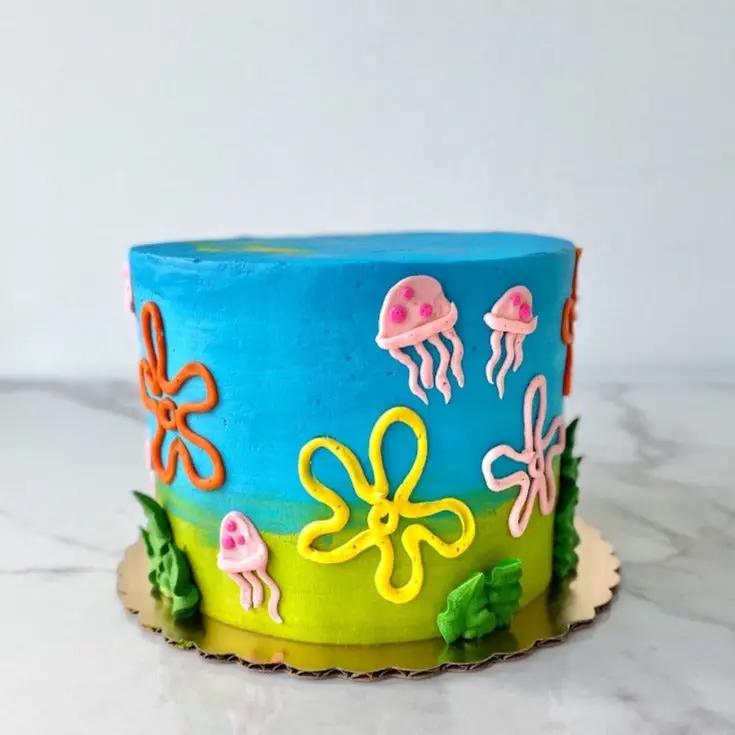 A round cake decorated to look like a SpongeBob SquarePants-themed cake. The cake has a light blue background with sea plant decorations, a large yellow, orange, and pink jellyfish.