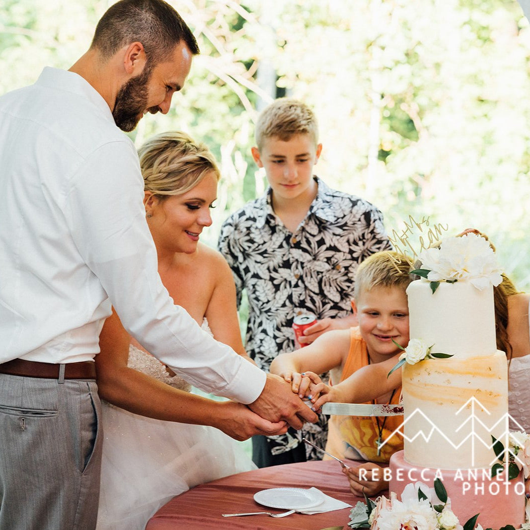 Groom cutting wedding cake with a large smile while bride watches joyfully, both surrounded by natural light and elegant table decor