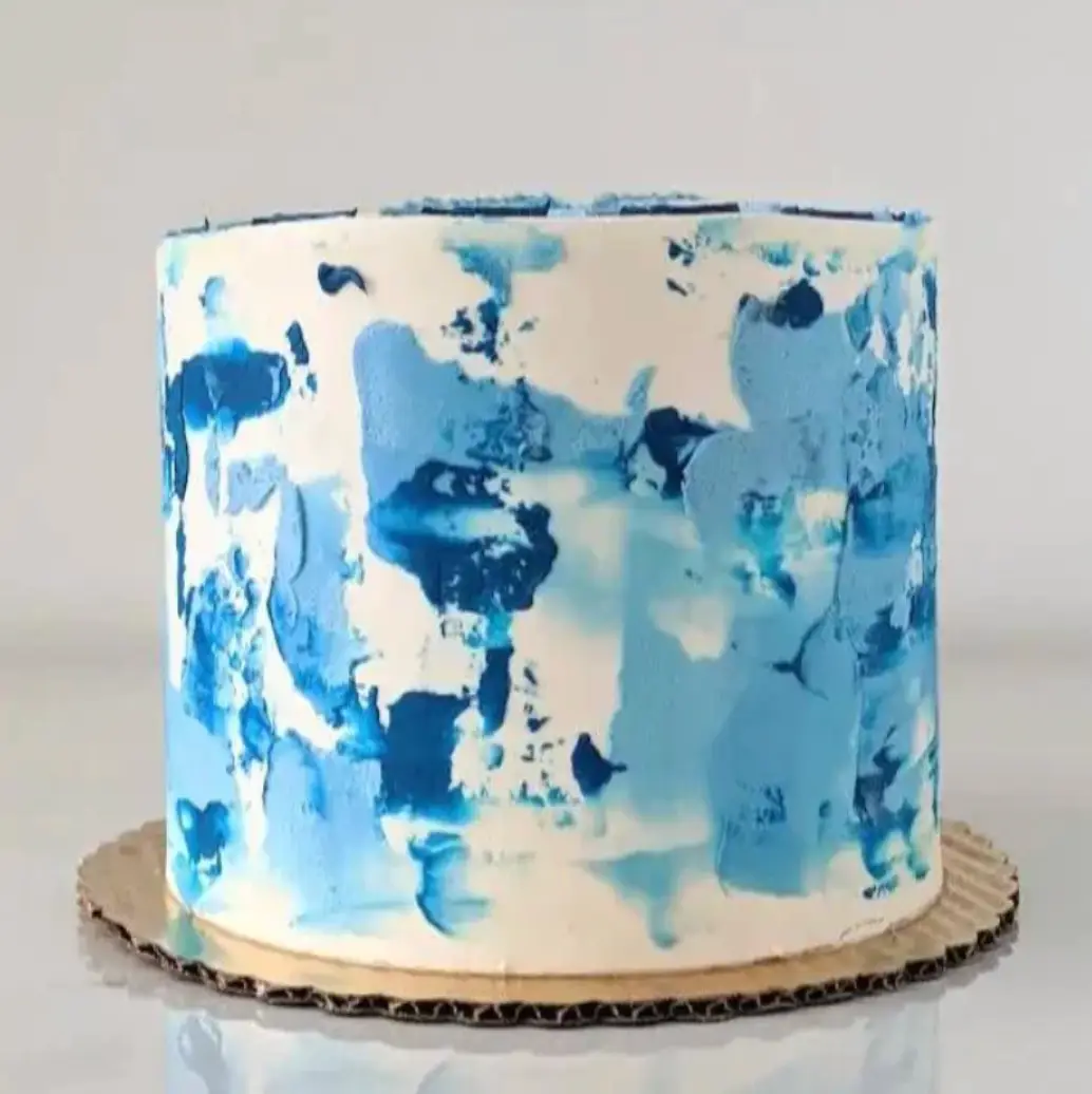 A round cake with white frosting and blue, abstract smears of icing on a gold cake board