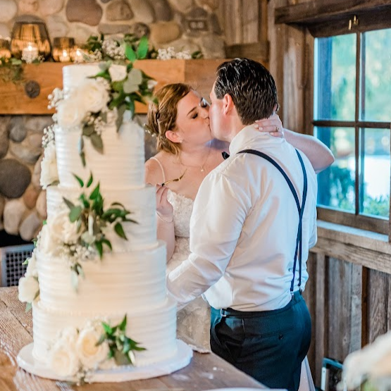 Baker in white apron carefully decorating a multi-tiered white wedding cake with fresh florals in a naturally lit room near large windows