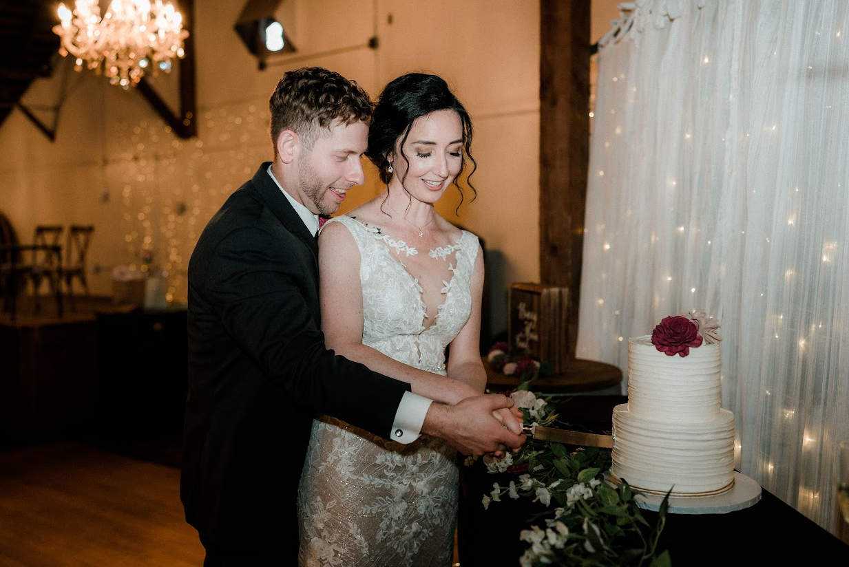 Bride and groom cutting their wedding cake indoors, surrounded by guests in a warmly lit reception hall