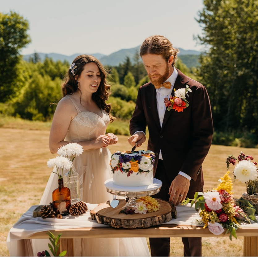 Outdoor wedding cake cutting scene with a bride and groom dressed in formalwear, smiling as they cut the cake together on a wooden table