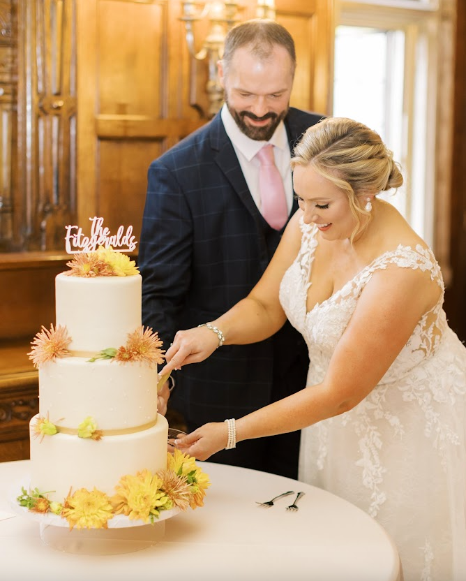 Happy bride and groom cutting a three-tier yellow and white wedding cake with smiles and joy during their indoor reception