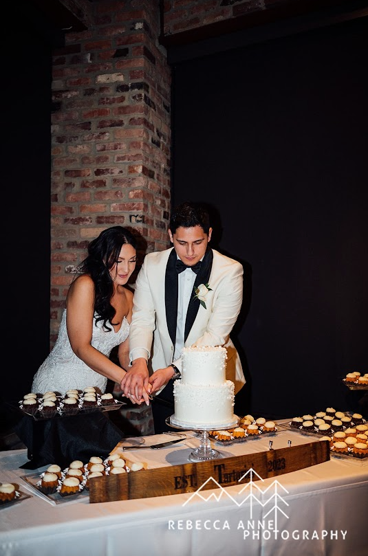 Romantic moment of a couple cutting a two-tier wedding cake on a rustic wooden table, captured in a dimly lit, cozy setting