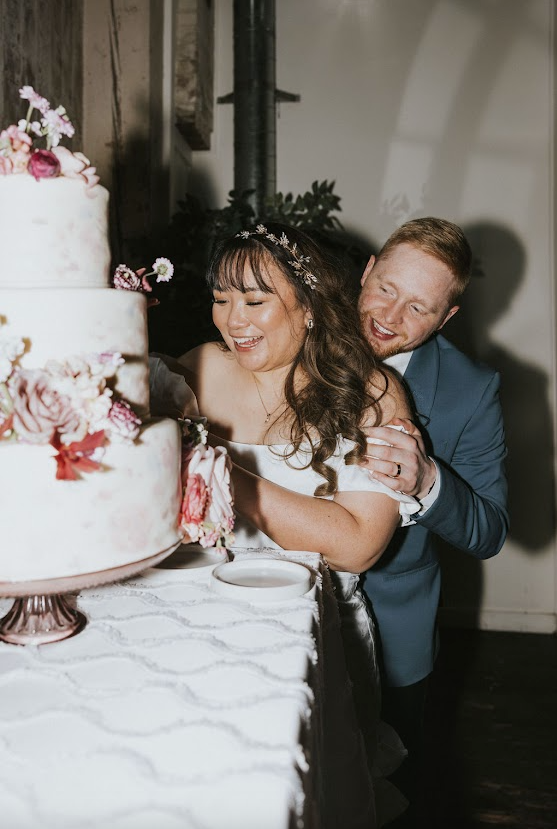 Newlywed couple sharing a playful cake moment, laughing as the groom offers the bride a bite during the wedding cake cutting ceremony