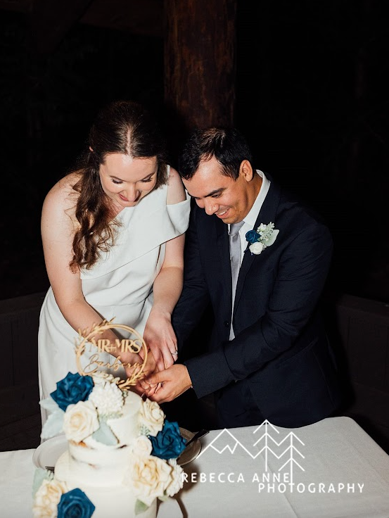 Bride and groom joyfully feeding each other wedding cake, surrounded by guests and celebration, both dressed in formal wedding attire