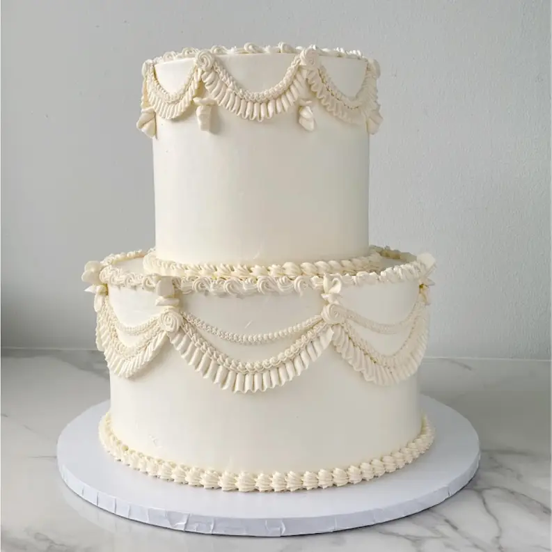 A two-tiered, white cake. Both tiers are decorated with elegant, rope-like frosting swirls and white lace trim.